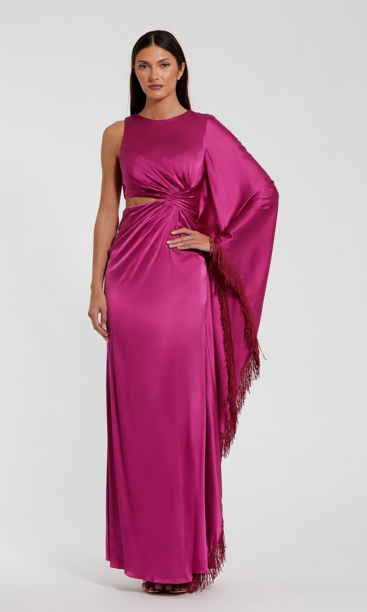 A woman stands against a plain background wearing the Mac Duggal 49898, a floor-length magenta satin formal dress featuring an asymmetrical sleeve, side cutout, and fringe detail.